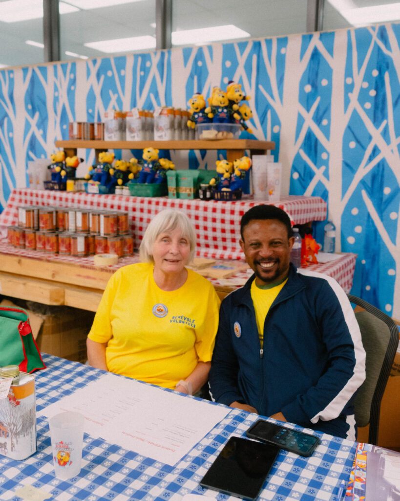 Smiling volunteers welcoming visitors at a market booth, inviting people to get involved in the Maple Sugar Festival.